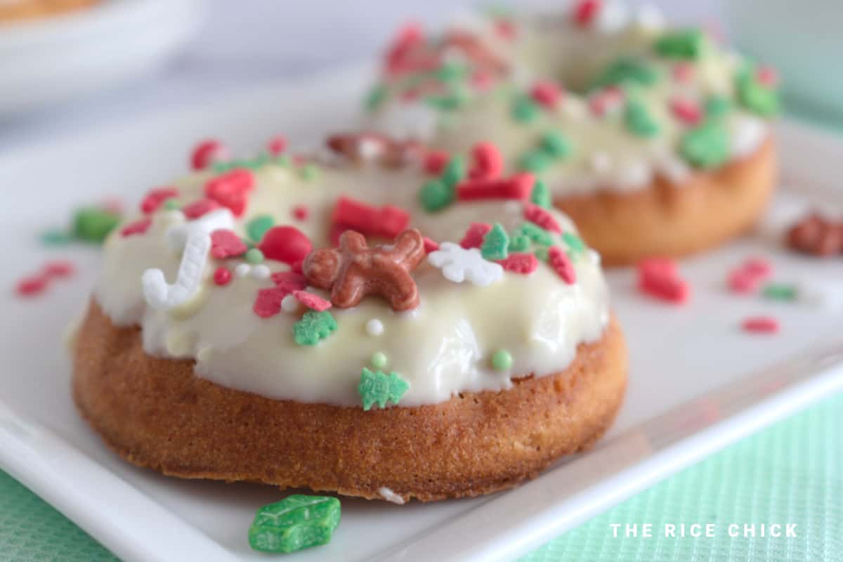 Two baked mochi donuts with a white chocolate glaze and Christmas sprinkles on a white rectangular plate.