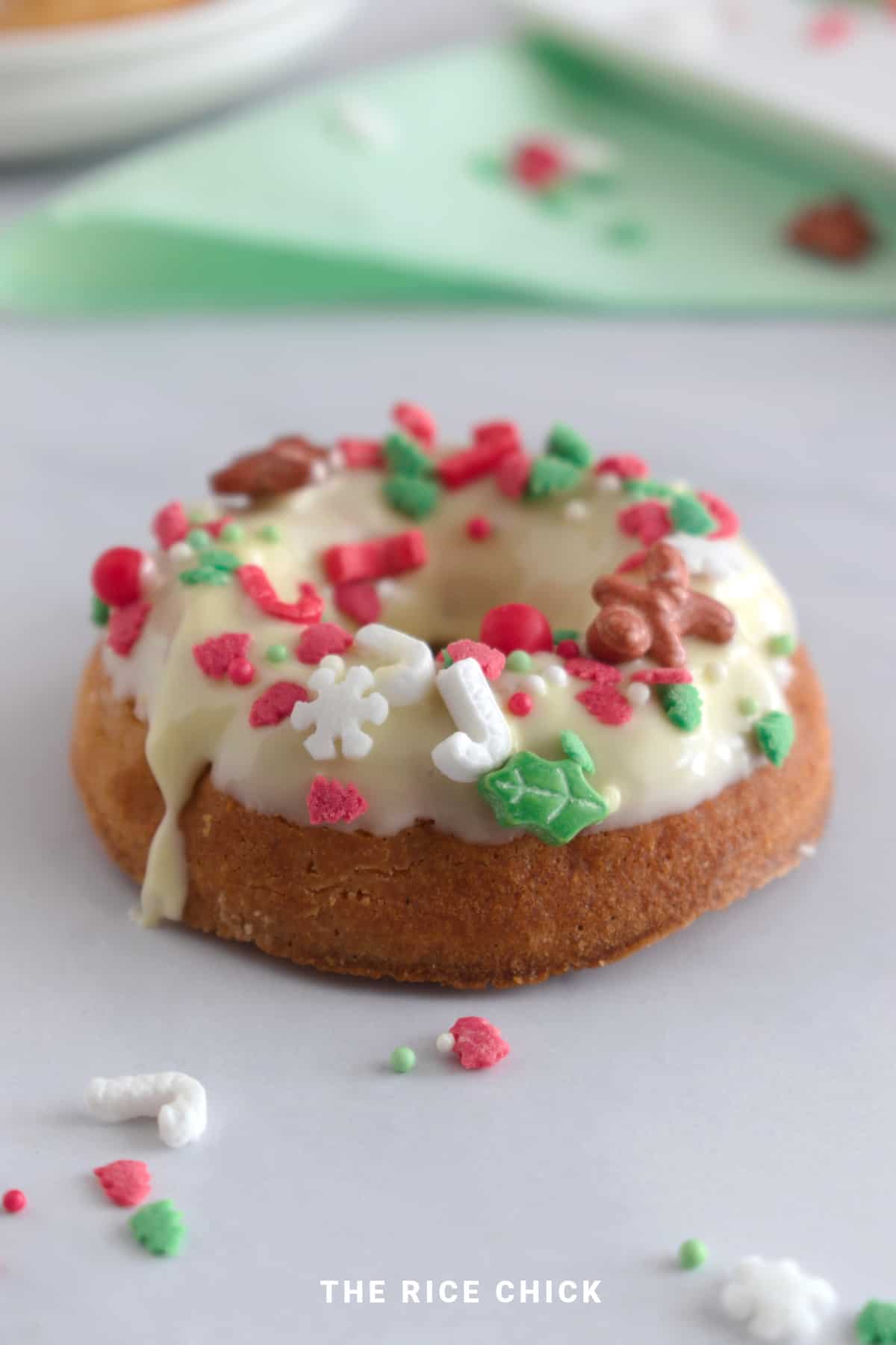 A baked mochi donut with white chocolate glaze and Christmas sprinkles on a white benchtop.