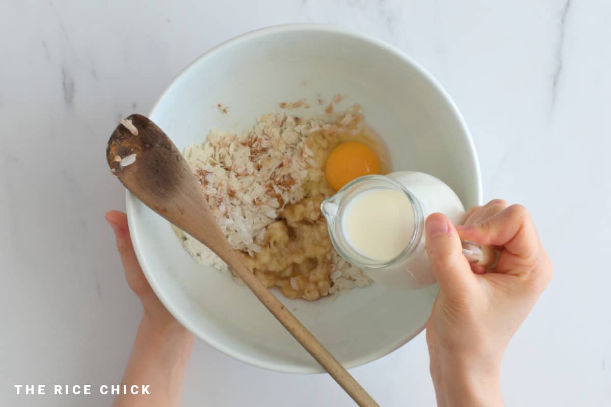 Pouring milk into a mixing bowl with rice flakes, ground cinnamon, banana, and egg.