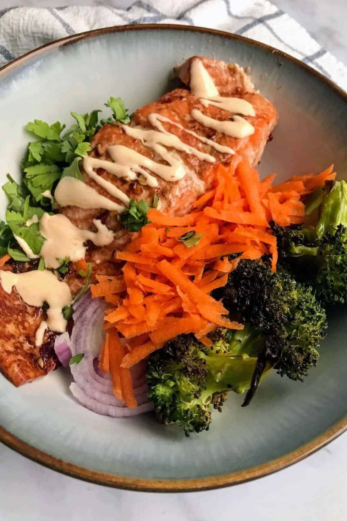 Grated carrot, broccoli, sliced red onion, salmon, and coriander leaves in a bowl.