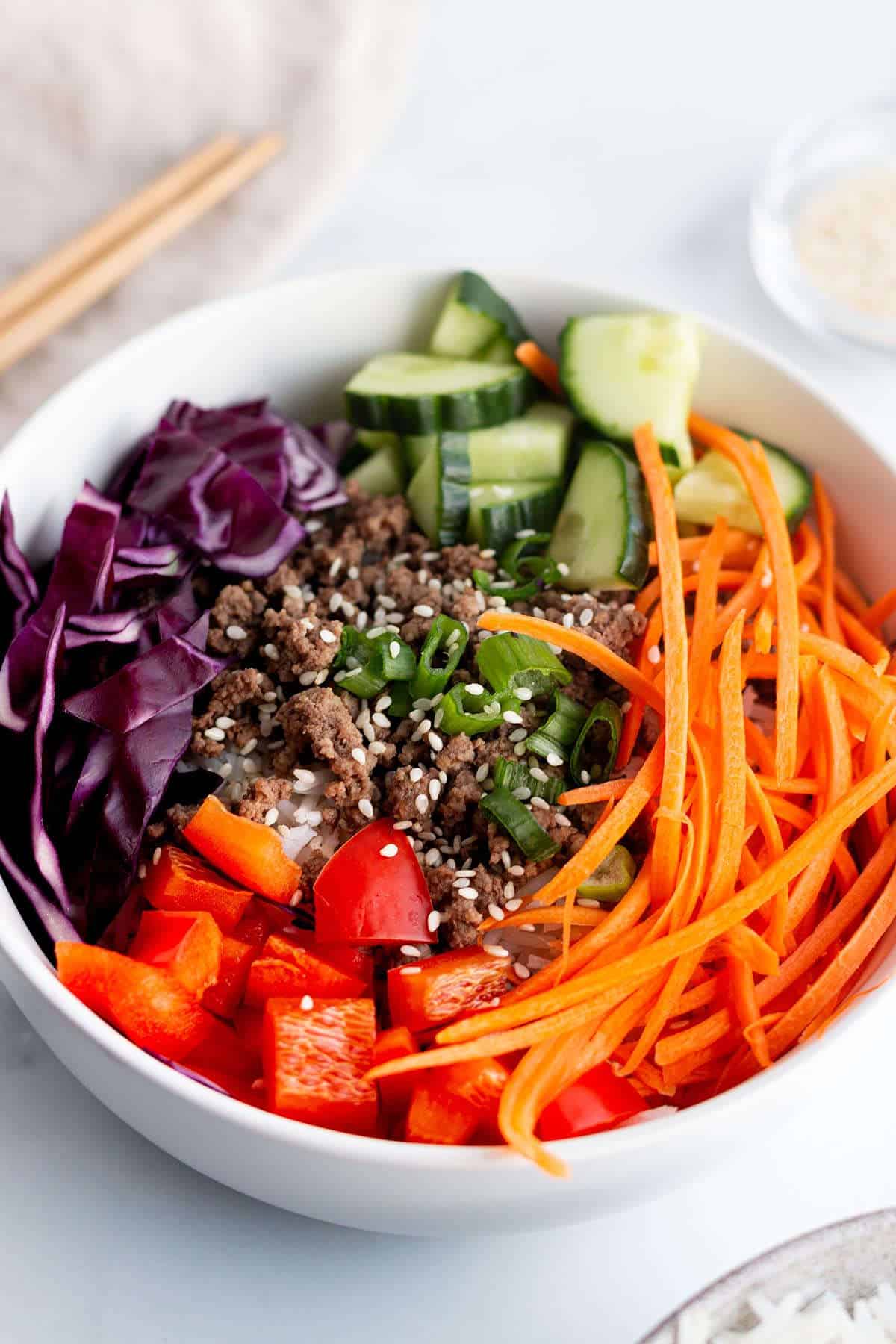 Ground beef, rice, shredded carrots, cucumber, cabbage, and green onions in a bowl.