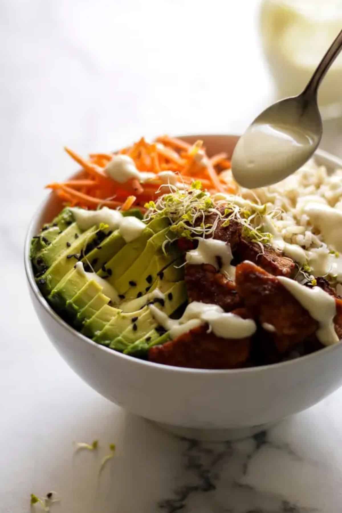 Spooning dressing over a rice bowl with chicken, avocado, rice, and carrots.