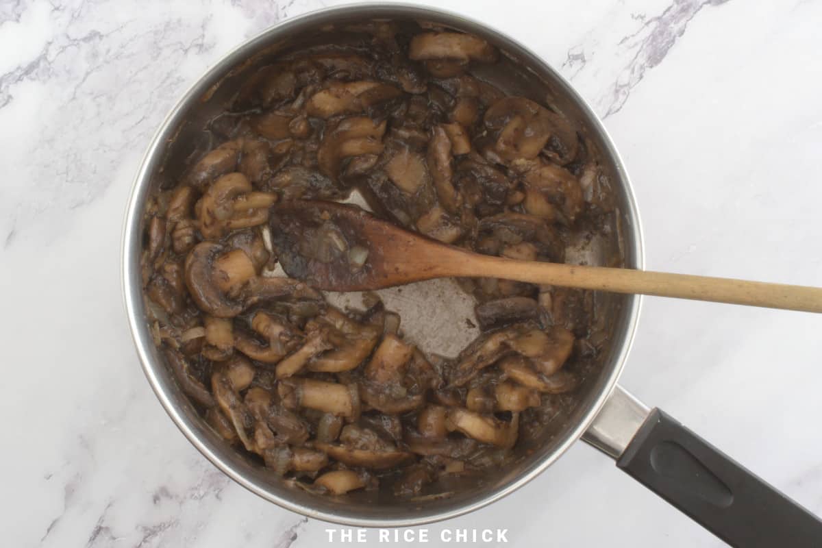 Cooked mushrooms and onions in a saucepan with a wooden spoon.
