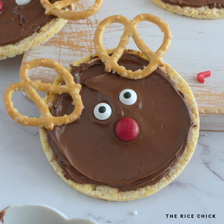 A reindeer rice cake with a red M&M, pretzels, Nutella, and candy eyes.