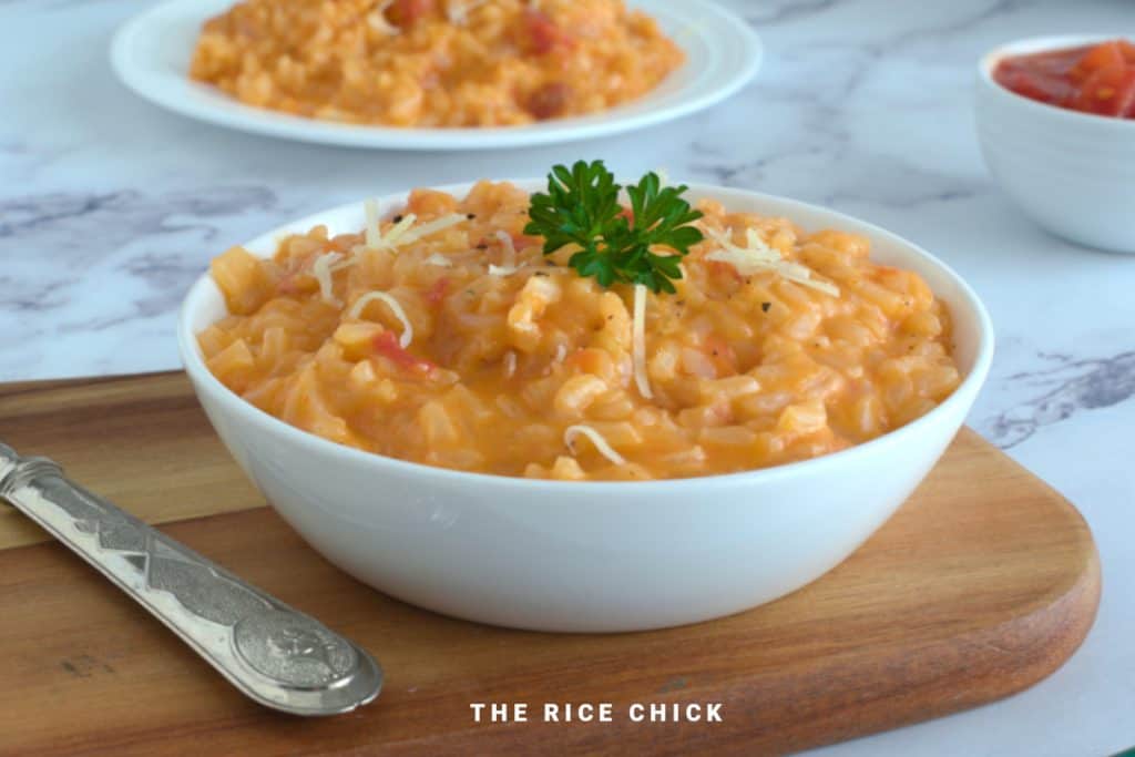 A white bowl filled with creamy risotto garnished with a sprig of parsley and shredded cheese sits on a wooden board with a spoon. Another bowl of risotto and a small bowl of sauce are in the background. The text "THE RICE CHICK" is at the bottom.