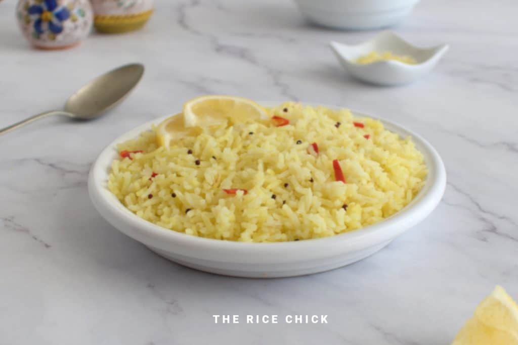 A white bowl filled with yellow seasoned rice garnished with lemon slices. The bowl is placed on a marble surface with a spoon and small decorative items in the background. Text at the bottom reads "THE RICE CHICK.